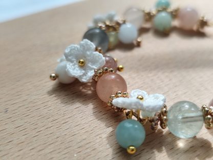 Handcrafted 'Aurelia Mist' micro crochet accessory featuring intricate, delicate patterns in soft pastel hues; an elegant, lightweight piece perfect for adding a touch of sophistication to any outfit. Displayed on a light-tone wood plate, under sunlight. The micro crochet white flowers are showing along side with the natural stone beads. The natural flourite stone beads are translucent and shining under the sunlight.
