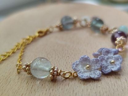 the elegant bracelet with hand dyed purple micro crochet flower with a golden beaded center and translucent crystal beads of purple, green and white color is shown. The bracelet laid on top of a light tone wooden board. The background is white.