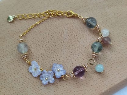 the elegant bracelet with hand dyed purple micro crochet flower with a golden beaded center and translucent crystal beads of purple, green and white color is shown. The bracelet laid on top of a light tone wooden board. The background is white.