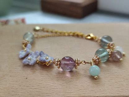 the elegant bracelet with hand dyed purple micro crochet flower with a golden beaded center and translucent crystal beads of purple, green and white color is shown. The bracelet laid on top of a light tone wooden board. The background is white.