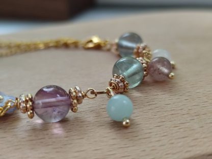 the elegant bracelet with hand dyed purple micro crochet flower with a golden beaded center and translucent crystal beads of purple, green and white color is shown. The bracelet laid on top of a light tone wooden board. The background is white.